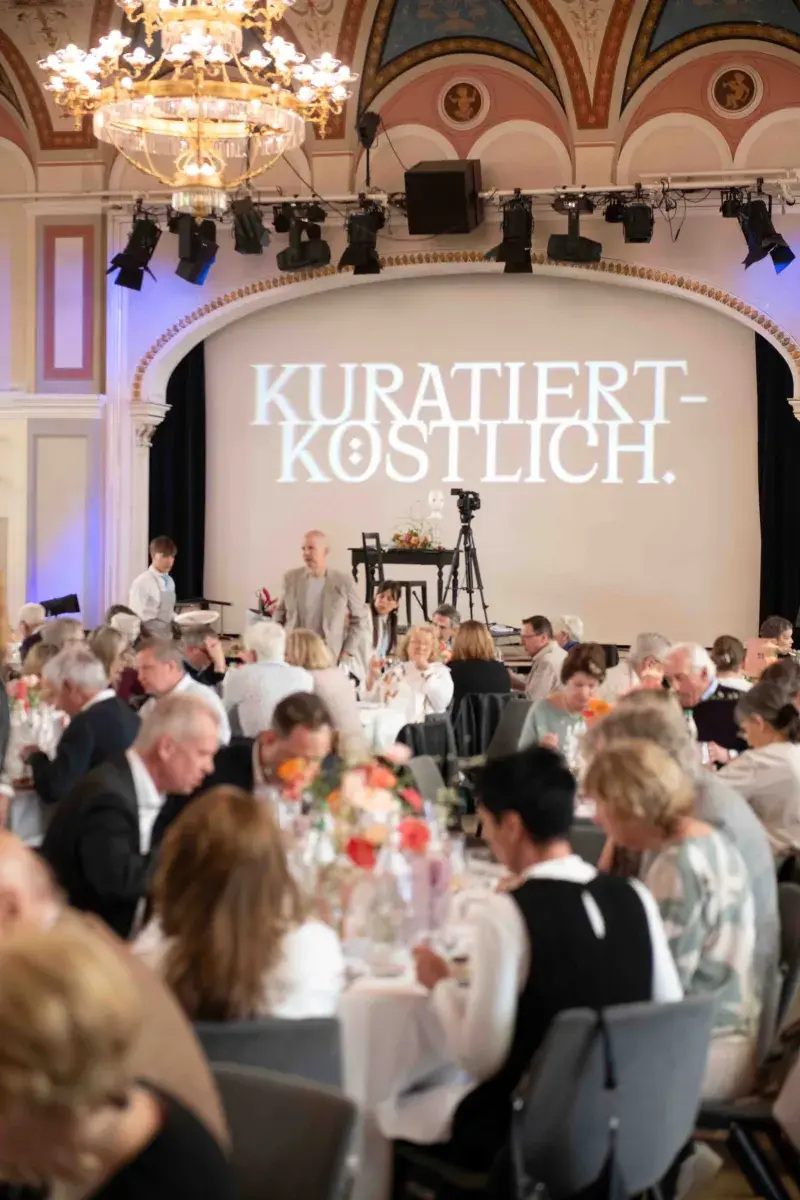 Publikum an den Tischen vor der Bühnenprojektion «Kuratiert Köstlich» im Kultursalon